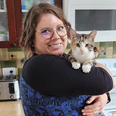 Picture of Marissa &mdash; the owner, trainer, &amp; ceramic artist behind Sunshine Paws &mdash; smiling while holding a mischievous cat in a brightly-lit kitchen