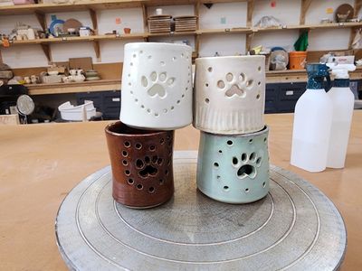 4 candle holder votives with paw print shapes hollowed out in them, stacked 2-by-2 in white, brown, & teal colours with a pottery studio in the background.